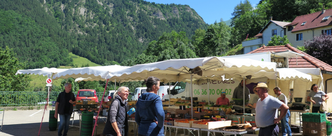 Photo avec un stand de fruits et lgumes, des personnes discutent en premier plan et il y a des montagnes en arrire-plan.