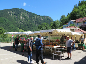 Photo avec un stand de fruits et lgumes, des personnes discutent en premier plan et il y a des montagnes en arrire-plan.